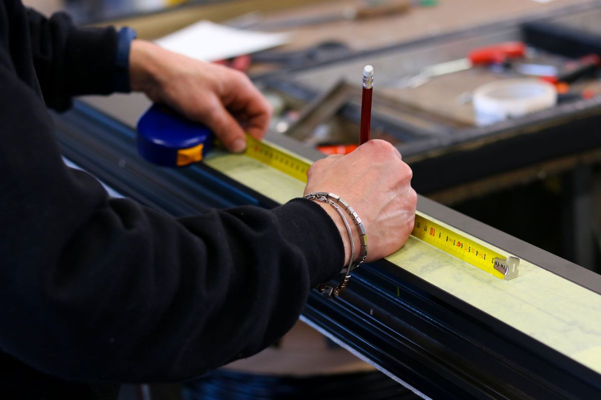 Hands measuring a dark aluminium profile with a tape measure and pencil on a workbench